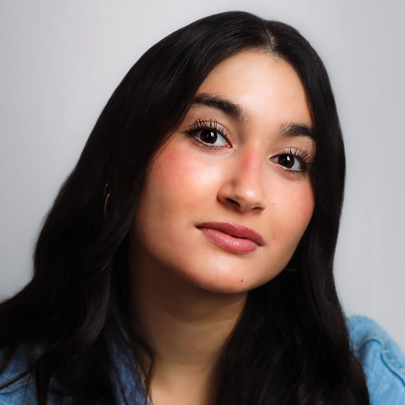 A young woman with long, dark wavy hair looks directly at the camera. She has natural makeup, rosy cheeks, and defined lashes. She is wearing a light denim jacket. The background is plain and light-colored, and the lighting is soft—perfectly capturing the effortlessly radiant style of Sara Ross Avery.