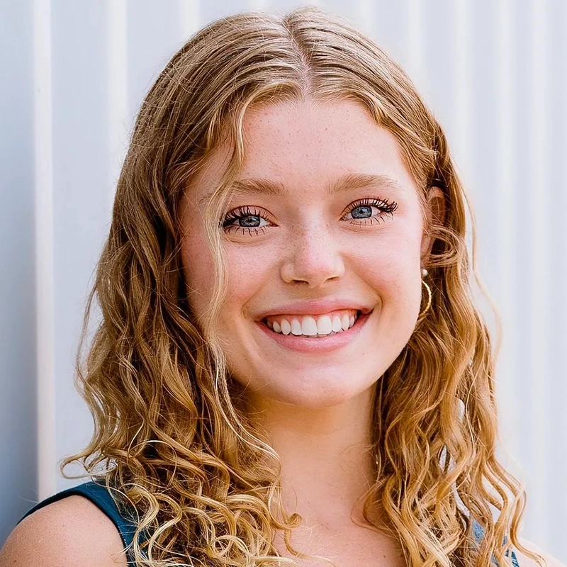 A young woman with long, curly blonde hair smiles warmly at the camera. She has blue eyes, light skin with freckles, and wears small hoop earrings. The background features a light-colored, vertical corrugated metal wall. Jordan Whitrock.