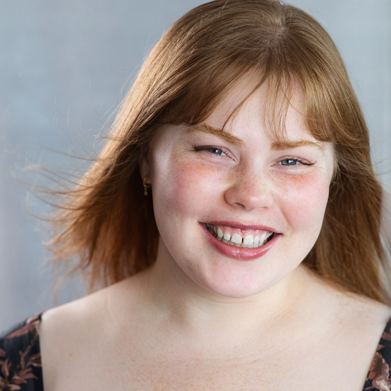 A young woman with fair skin and straight, light brown hair smiles warmly. Roni Ellison has freckles and is wearing a dark floral top. The background is softly blurred in gray tones, her hair gently blown back—she appears happy and relaxed.