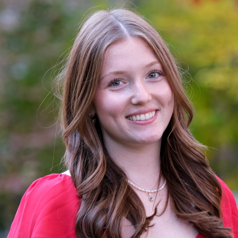 A young woman with long, wavy brown hair smiles at the camera. She is wearing a red top and layered pearl necklaces. The background is blurred greenery, suggesting an outdoor setting with natural light.