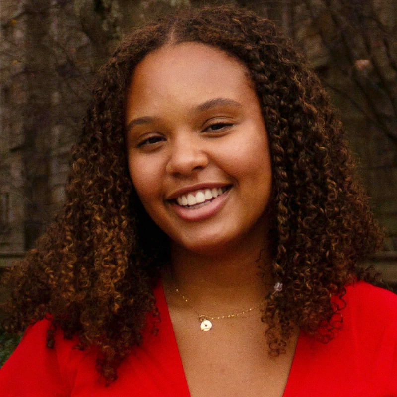 A young woman with medium-brown skin and curly brown hair smiles at the camera. She wears a red top and a delicate gold necklace with a small round pendant. The background is blurred, showing bare trees and hints of a building.