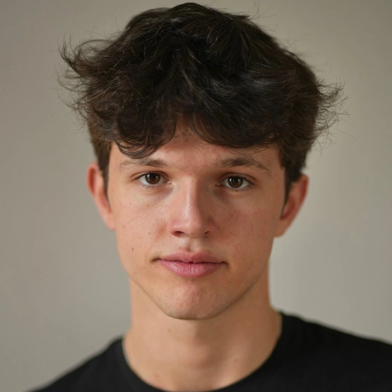 A young man with tousled dark brown hair and fair skin looks directly at the camera with a neutral expression. He is wearing a black shirt and is posed against a plain, light-colored background.