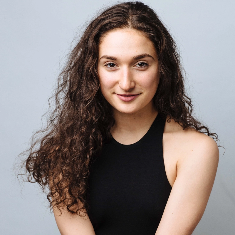A young woman with long, curly brown hair and fair skin smiles gently. Victoria Fiore wears a sleeveless black top and sits against a plain light gray background, looking directly at the camera with a relaxed, confident expression.