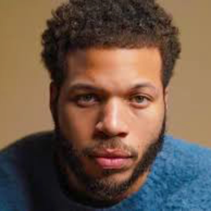 Mister Fitzgerald, a man with short curly hair and a full beard, looks directly at the camera. He wears a textured blue sweater and stands against a plain light brown background, with soft lighting highlighting his features and expression.