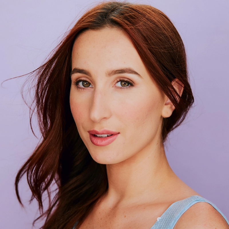 Madeleine Flanagan, a young woman with long, reddish-brown hair, poses in front of a light purple background. She has fair skin, light makeup, and wears a blue sleeveless top. Her expression is calm and confident, with hair gently draped over one shoulder.