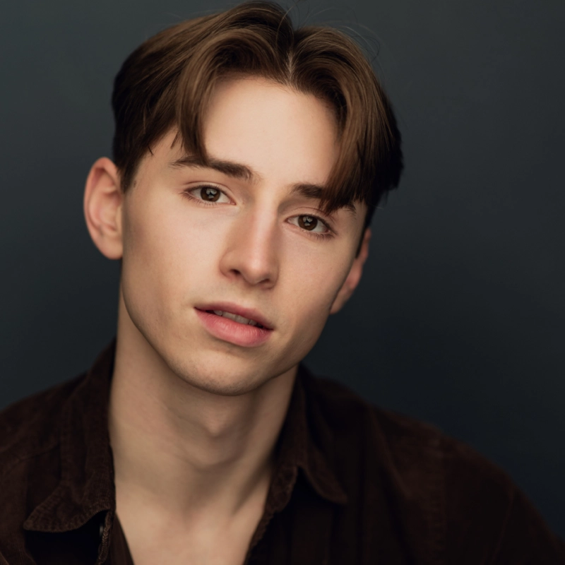 A young person with fair skin, straight brown hair parted down the middle, and brown eyes gazes softly at the camera. Wearing a dark collared shirt, Will Forrest is set against a dark background, soft lighting highlighting their facial features.