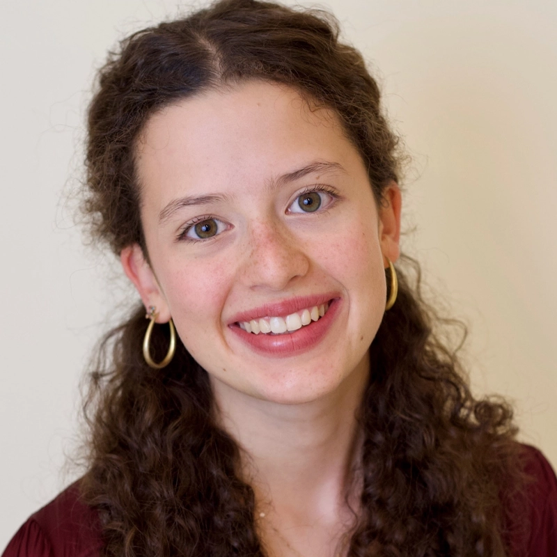 A young woman with long, curly brown hair and light skin smiles at the camera. She is wearing gold hoop earrings and a burgundy top. The background is plain and light-colored, keeping the focus on her friendly, expressive face.