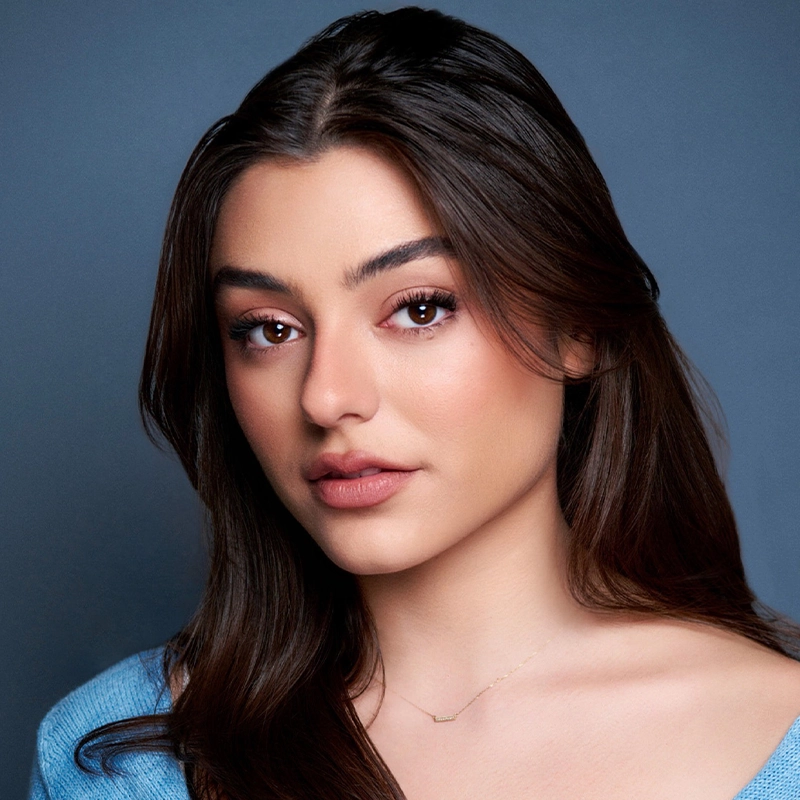 A young woman with long brown hair and brown eyes looks at the camera with a neutral expression. Alex Humphreys wears natural makeup, a soft blue top, and a delicate gold necklace. The background is a plain, muted blue-gray color.