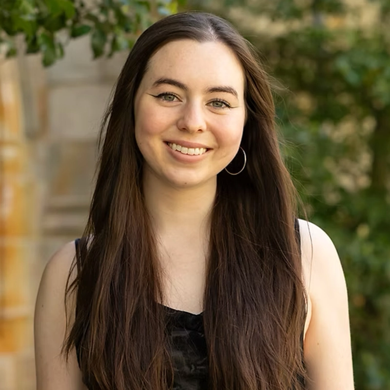 A young woman with long brown hair and hoop earrings smiles at the camera. She is wearing a sleeveless black top and stands outdoors, with greenery and a blurred stone wall in the background. The lighting is bright and natural.