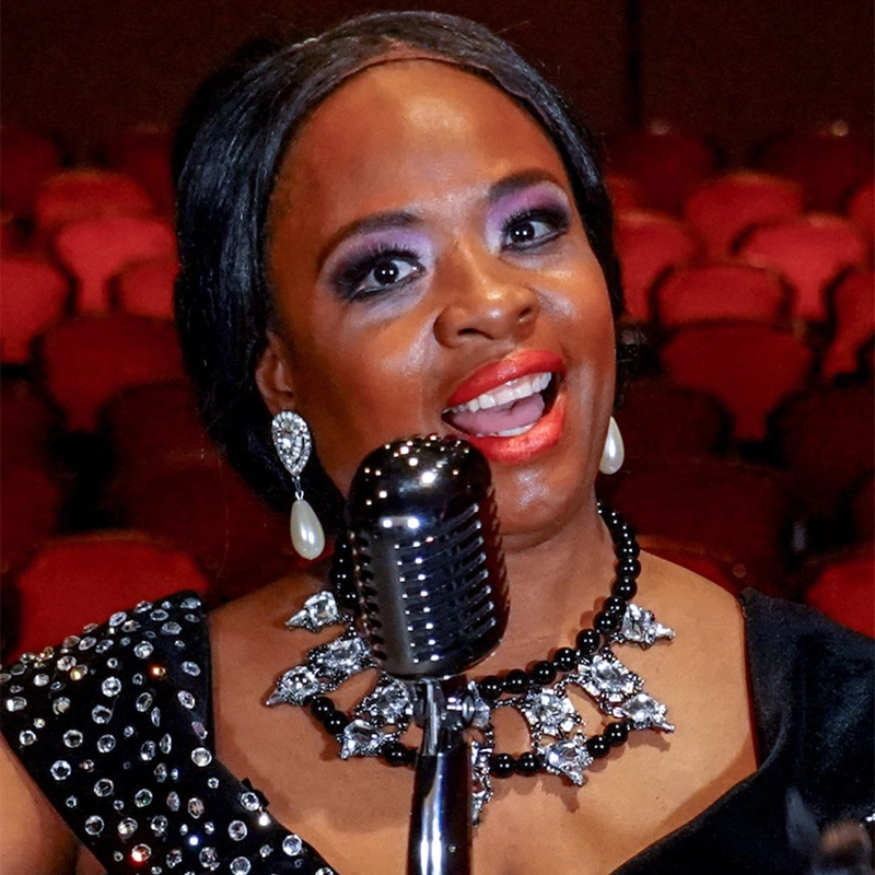 Anissa Gathers, in a black, jeweled dress, sings into a vintage microphone. She wears dramatic makeup, pearl earrings, and a sparkling necklace. Red theater seats blur in the background, highlighting the performance setting.