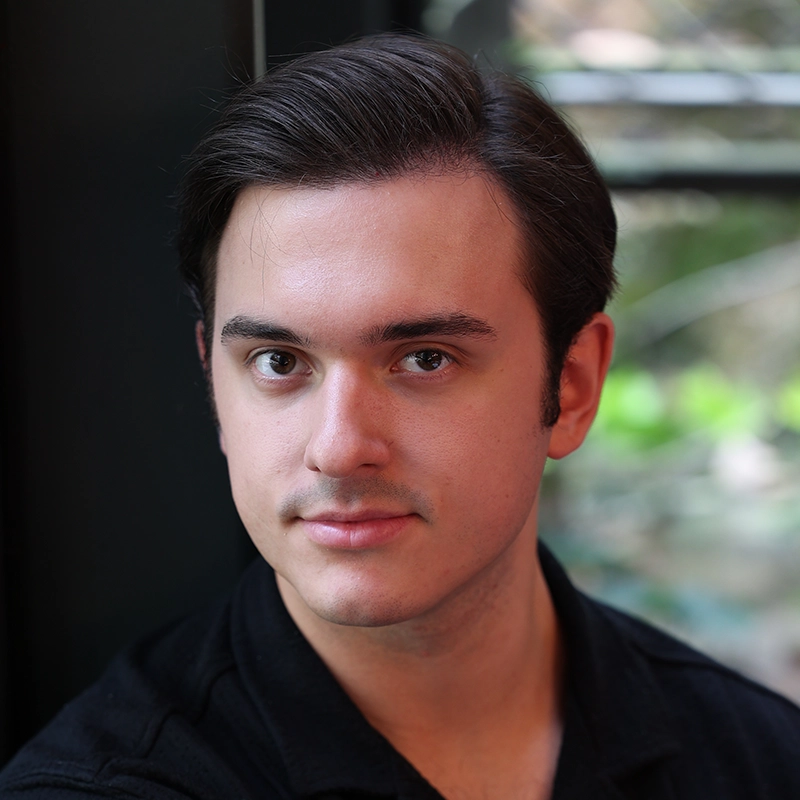 A young man with fair skin and dark brown hair, parted to the side, looks at the camera with a neutral expression. He wears a black collared shirt and sits indoors near a window with soft natural light and greenery visible outside.