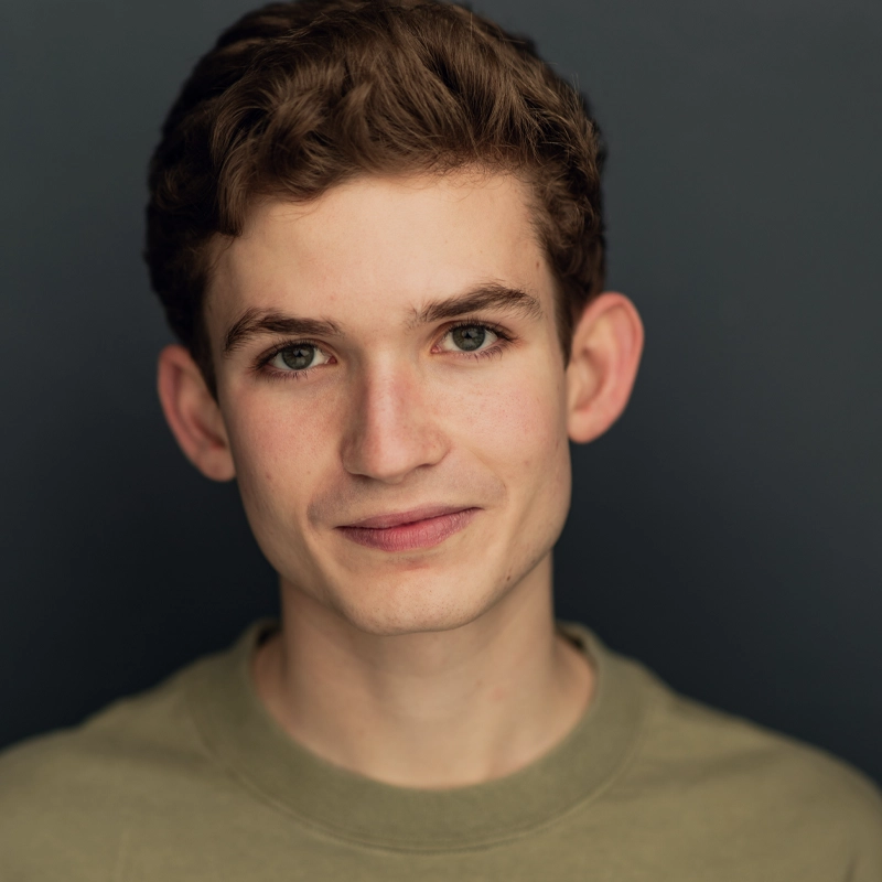 Luke Grayson, a young man with short, wavy brown hair and green eyes, smiles softly at the camera. He wears an olive green crewneck shirt against a plain dark background, with even lighting highlighting his facial features.