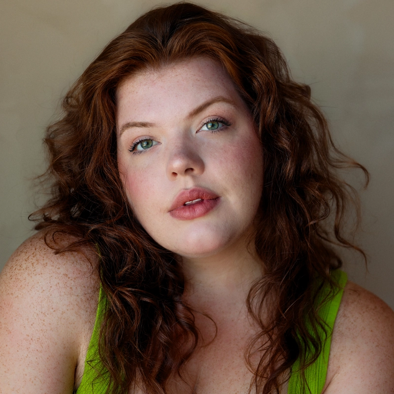 Shelby Griswold, with wavy auburn hair and fair, freckled skin, gazes softly at the camera. She has light blue-green eyes, natural makeup, and slightly parted lips. She wears a bright green sleeveless top against a neutral background.
