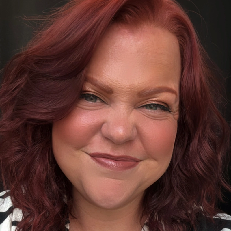 A woman with shoulder-length wavy auburn hair smiles warmly. She has fair skin, blue eyes, and is wearing natural makeup with rosy cheeks and lipstick. She is dressed in a black and white top and is posed against a dark background.