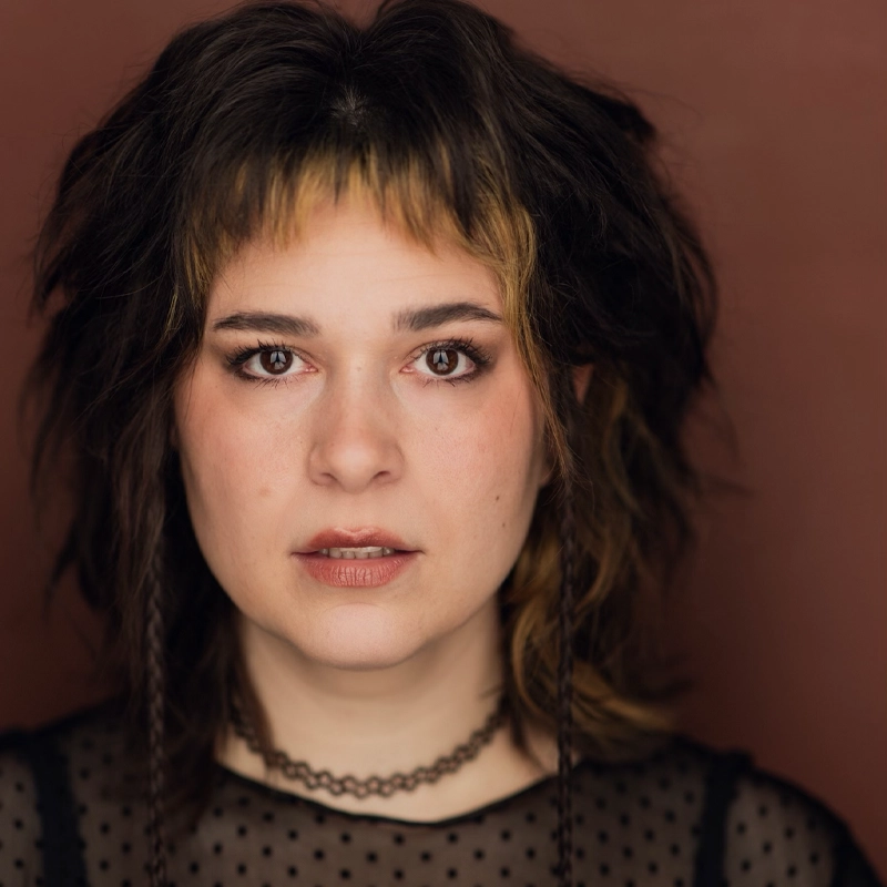 A person with layered, shaggy dark hair and blonde streaks faces forward against a brown background. They wear a sheer black polka-dot top and a choker necklace. Their expression is neutral, and their makeup includes eyeliner and natural-toned lipstick.