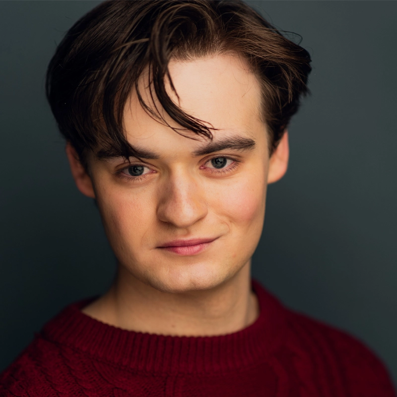A young man with fair skin and medium brown hair smiles softly. He wears a burgundy knit sweater and poses against a dark, plain background. His hair is parted to the side with loose strands falling onto his forehead.