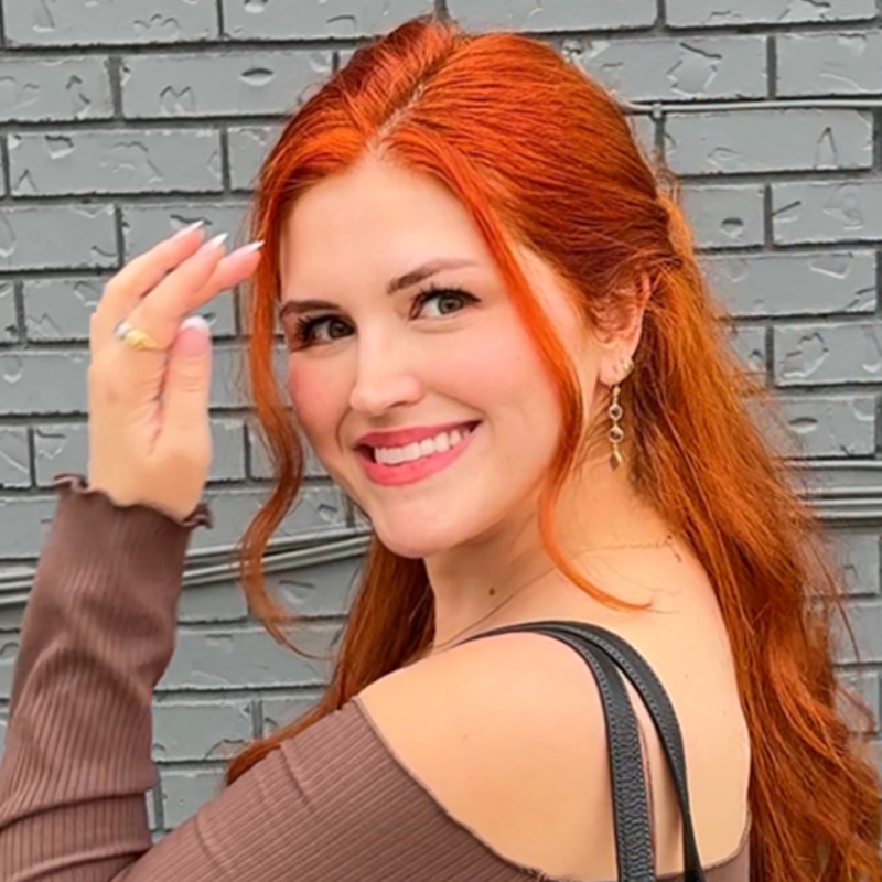 A woman with long, bright red hair smiles at the camera. She wears a brown off-shoulder top and dangling earrings, holding her hand near her face. In the background, there is a gray painted brick wall.
