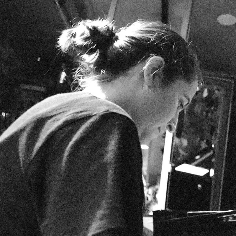 A black-and-white photo shows Hannah Mayer with long hair tied in a messy bun, wearing a dark T-shirt, viewed in profile. She is indoors, focused on something in front of her, with dim light and blurred background details.