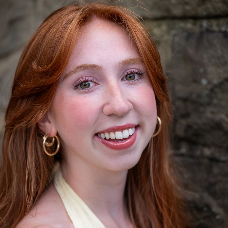 Emily Harajda, a young woman with long red hair and fair skin, smiles warmly. She wears gold hoop earrings and a sleeveless cream top. Her green eyes and minimal makeup highlight her natural beauty against the blurred stone background.