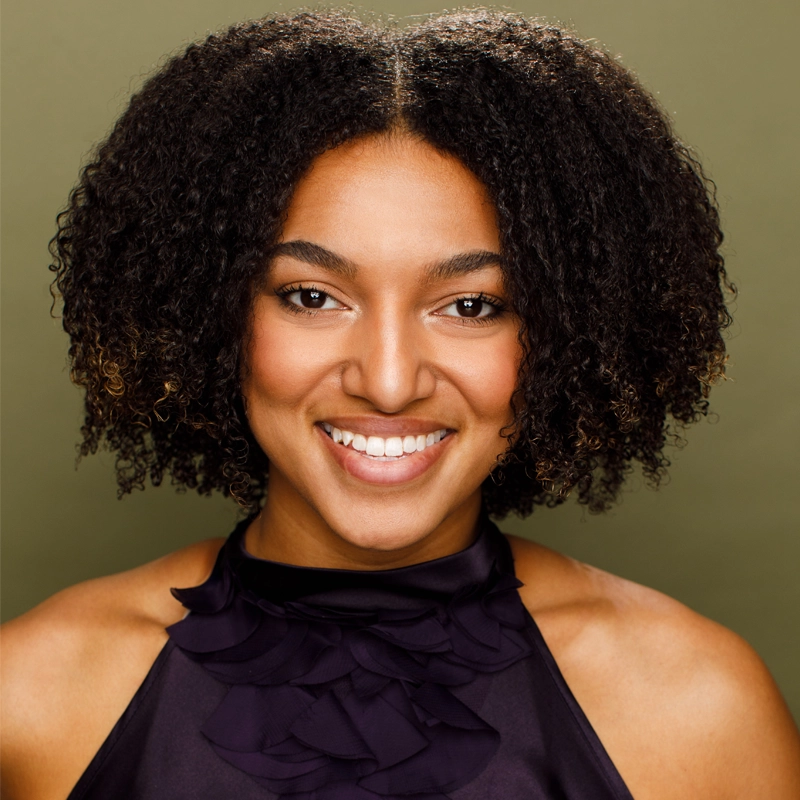 Anne-Sophie Hill, a young woman with medium brown skin and natural, curly dark hair, smiles warmly at the camera. She wears a sleeveless, ruffled black top against a plain olive-green background, with soft lighting highlighting her features.