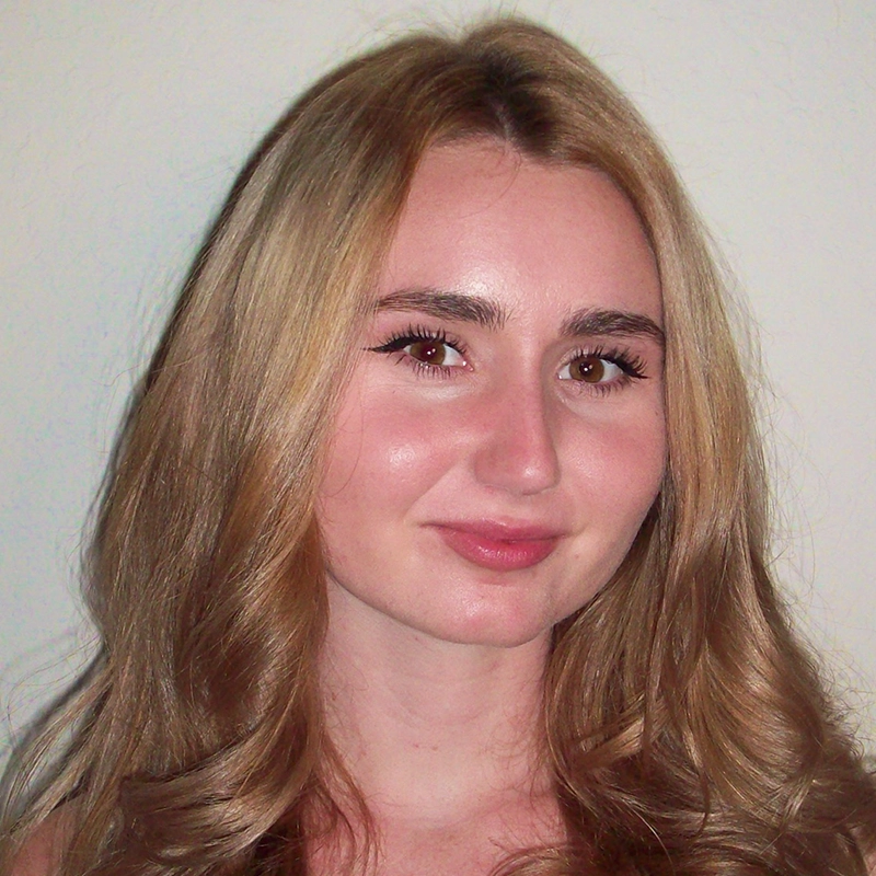 A young woman with long, wavy, blonde hair and fair skin is posing in front of a plain, light-colored background. She has full eyebrows, brown eyes, subtle makeup, and is giving a gentle, closed-mouth smile while looking directly at the camera.