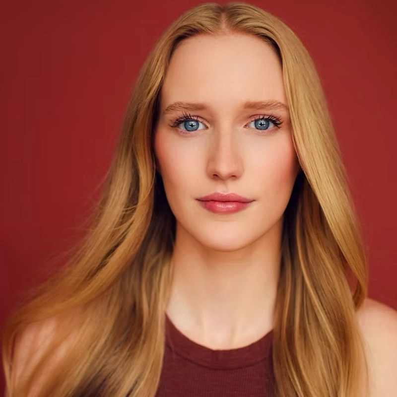A young woman with long, straight blonde hair and blue eyes gazes directly at the camera. She wears a sleeveless maroon top. The background is solid red, creating a warm, simple backdrop. Her expression is calm and confident.