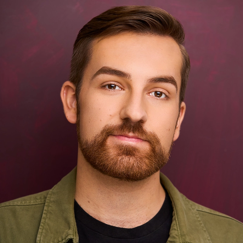 A young man with light skin, neatly styled brown hair, and a trimmed beard looks into the camera with a slight smile. He is wearing a green jacket over a black shirt. The background is a solid, textured maroon color.