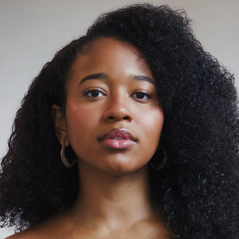 A young woman with medium brown skin and voluminous, curly black hair looks directly at the camera. She wears hoop earrings and has a calm, neutral expression. The background is plain and softly lit.