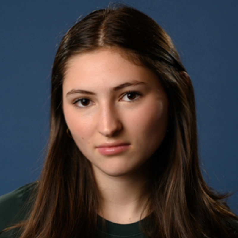 A young woman with long brown hair and fair skin, wearing a dark green top, looks directly at the camera with a neutral expression. The solid blue background brings focus to Micah Wolfensohn's poised presence.