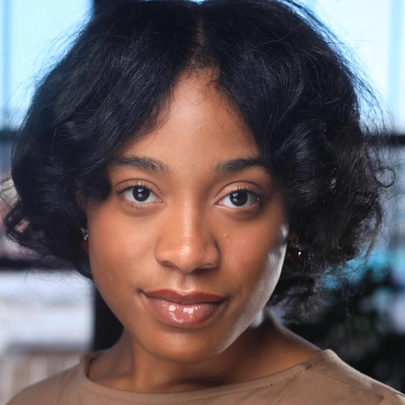 A young woman with medium-brown skin and short, wavy black hair looks directly at the camera. She wears a light brown top and subtle makeup. The background is softly blurred with natural light coming through large windows.
