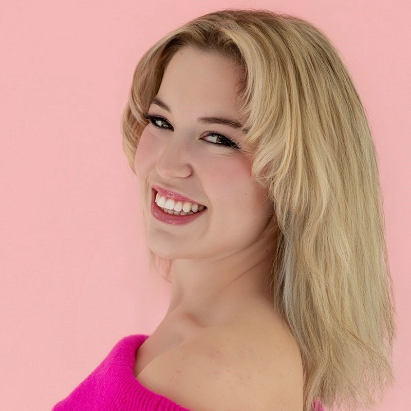 A young woman with shoulder-length blonde hair smiles brightly, looking over her shoulder. She wears a vibrant pink off-the-shoulder top and stands against a soft pink background, giving the image a cheerful and upbeat atmosphere.
