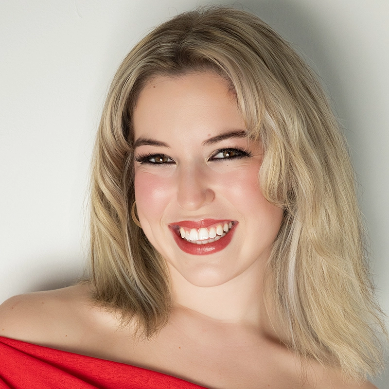A woman with shoulder-length blonde hair smiles brightly. She has fair skin, is wearing glossy red lipstick, and has light makeup. She wears a red off-shoulder top and stands against a plain light background.