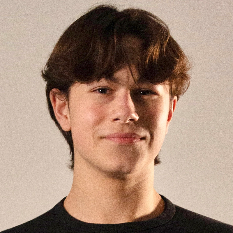A teenage boy with medium-length brown hair and fair skin smiles slightly. Micah Wolfensohn wears a black crewneck shirt and stands against a plain, light gray background. The lighting is soft, and his expression is calm and relaxed.