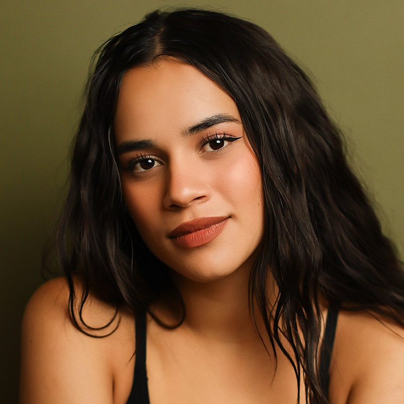 Lexis Trechak, a young woman with long, wavy dark hair and warm skin tone, looks at the camera in a black tank top. Her subtle makeup in natural tones stands out against the solid olive green background, creating a calm, neutral setting.