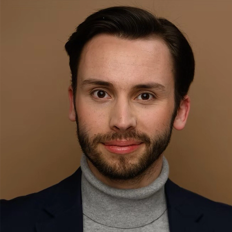 Jordan Whitrock, a young man with neatly styled dark hair and a short beard, is smiling slightly. He wears a gray turtleneck and a navy blazer. The plain, warm brown background and even lighting highlight his features.