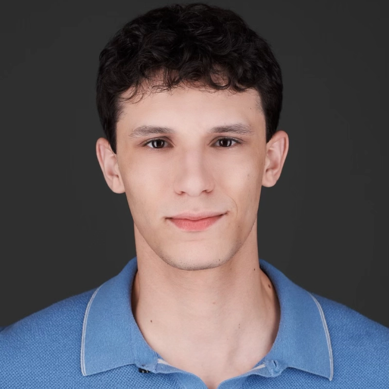 A young man, Eli Jacobson, with short, curly dark hair and fair skin faces the camera with a calm expression. He wears a light blue collared shirt against a plain, dark gray background. The even lighting highlights his facial features clearly.