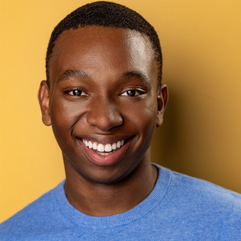 A young man with short hair smiles warmly at the camera. He is wearing a light blue crewneck shirt and stands against a solid mustard yellow background. The lighting is bright and even, highlighting his joyful expression.