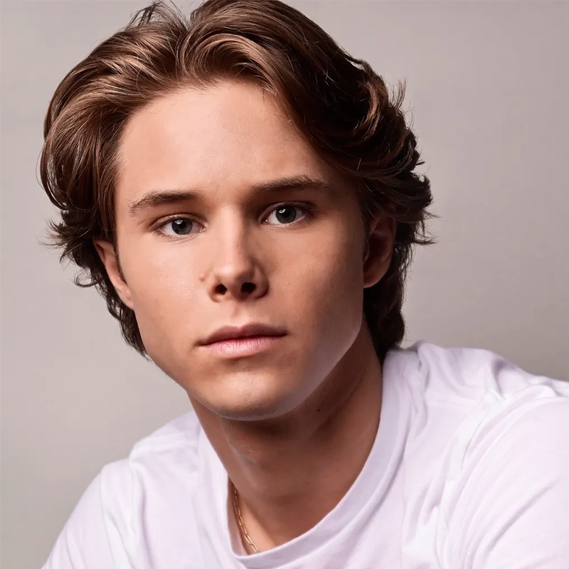A young person with light skin, brown eyes, and medium-length, light brown hair combed back, wearing a white t-shirt, looks directly at the camera with a neutral expression—resembling Violet Tinnirello—against a plain, light-colored background.