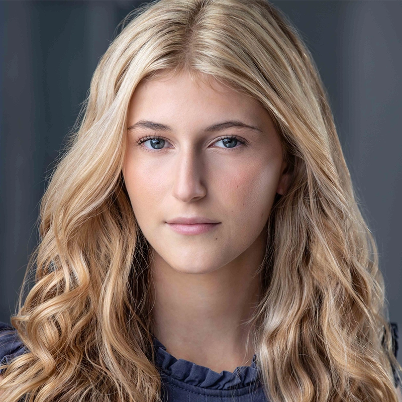 A young woman with long, wavy blonde hair and fair skin looks directly at the camera with a neutral expression. She is wearing a dark ruffled top, and the background is softly blurred in shades of dark gray and blue.