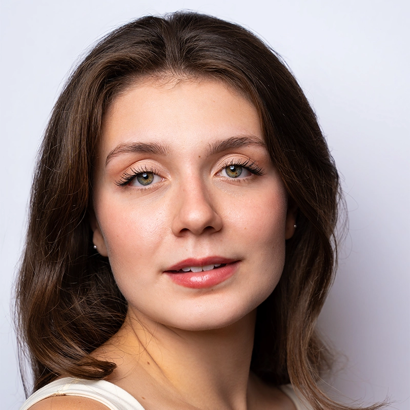 Jenna Willet, a young woman with fair skin and long brown hair, looks at the camera with a slight smile. She has light hazel eyes, natural makeup, and wears a white top against a softly lit, plain background that highlights her clear complexion.