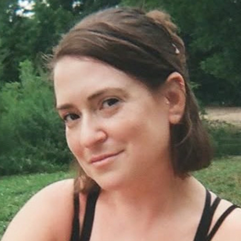 Jess Honovich, a woman with light skin and a short brown bob, sits outdoors amid greenery. She wears a black sleeveless top and offers a gentle, slight smile to the camera in natural lighting.