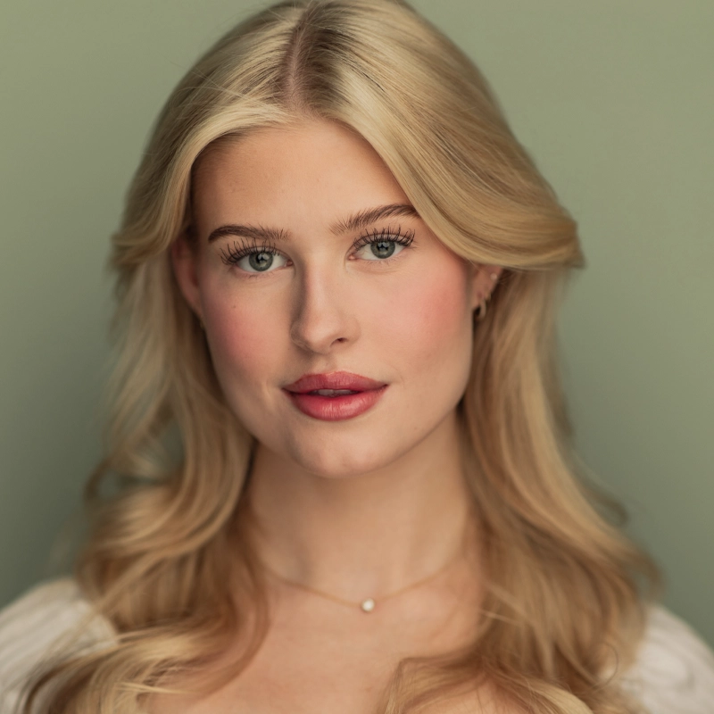 Charlotte Jensen, a young woman with long, wavy blonde hair and blue eyes, looks directly at the camera. She has fair skin, natural makeup with pink lips and flushed cheeks, and wears a delicate necklace. The background is a soft, muted green.
