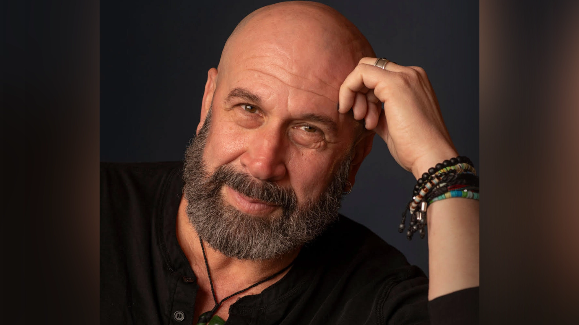 Jeremy Schonfeld - Site Image A bald, bearded man with a friendly expression rests his head on his hand. He wears a black shirt and multiple bracelets, including beaded and leather bands. The dark background draws focus to Jeremy Schonfeld, known for his heartfelt songs.
