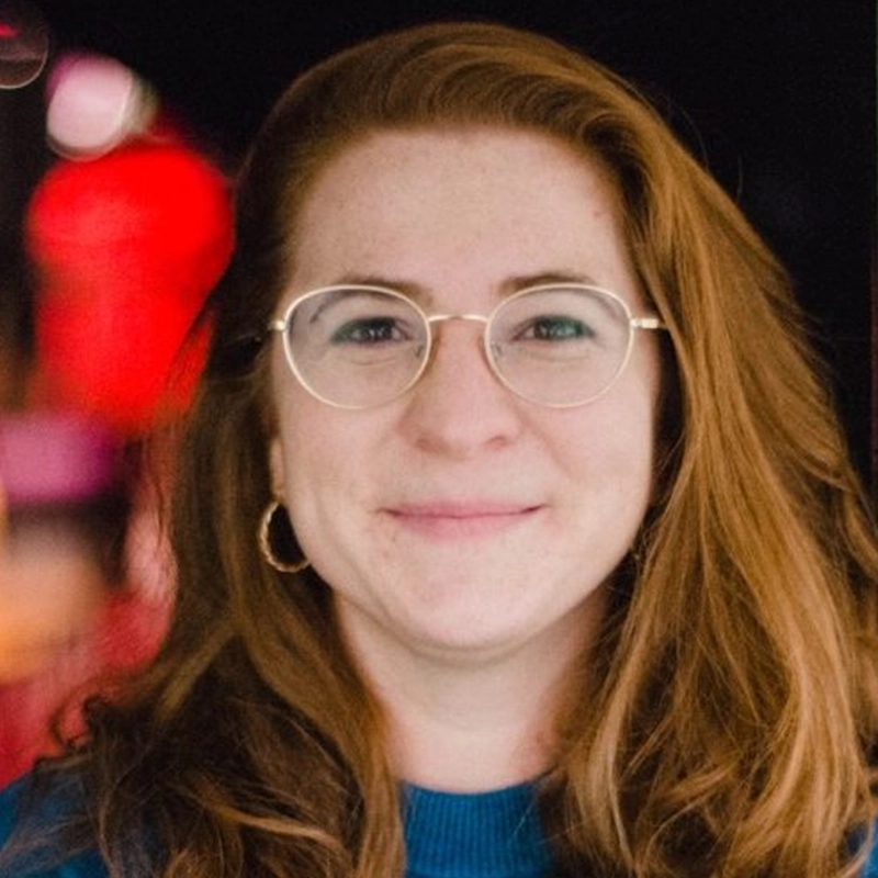 Jess Honovich, with long, wavy red hair, smiles gently. She wears round, silver-rimmed glasses, a blue top, and a gold hoop earring. The out-of-focus background glows with colorful lights, creating a warm, soft ambiance.