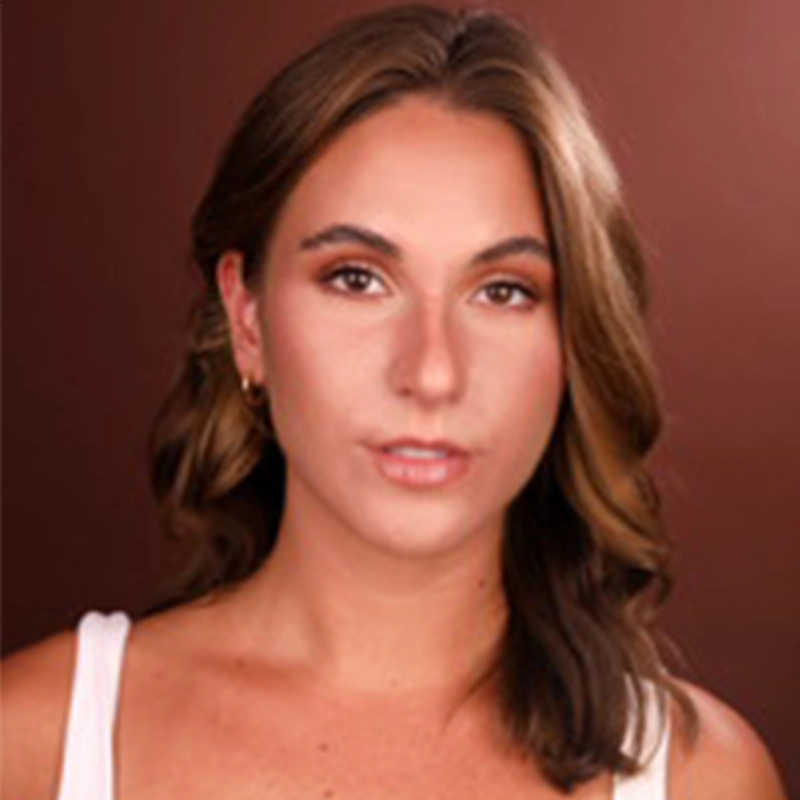 A young woman with light skin and wavy brown hair looks directly at the camera. She wears a white top and small hoop earrings. The background is a solid brown color, and her expression is calm and neutral.