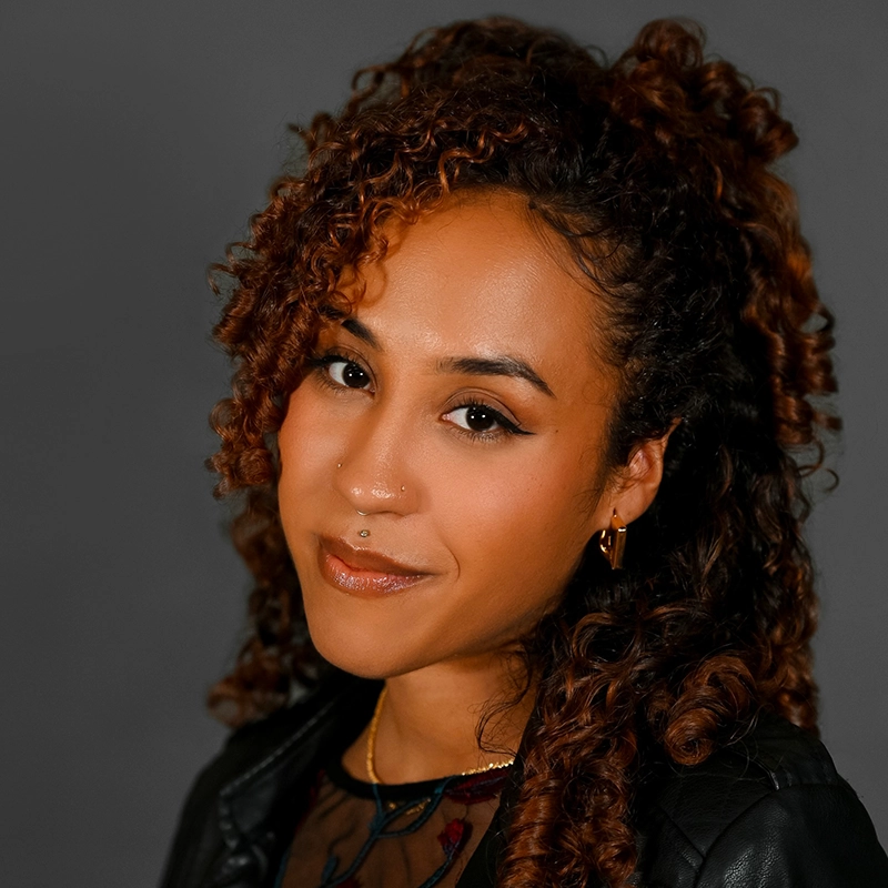 Jenna Willet, with curly auburn hair styled half-up, poses against a gray background. She has warm brown skin, winged eyeliner, a nose piercing, hoop earrings, a gold necklace, sheer black top, and dark jacket—smiling softly at the camera.
