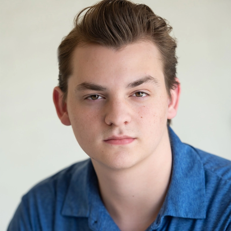 Zach Austin Johnson, a young man with fair skin and short, slicked-back brown hair, looks directly at the camera with a neutral expression. He is wearing a blue collared shirt and is posed against a plain, light-colored background.