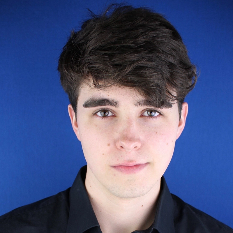 Jordan Whitrock, a young man with fair skin, short dark brown hair, and hazel eyes, looks directly at the camera with a neutral expression. He is wearing a black collared shirt and is posed against a solid blue background.