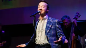 A young man, Josh Thone, sings into a microphone on stage, smiling and wearing a blue patterned blazer over a white shirt. A musician in the background plays an upright bass, with stage lights casting purple and blue tones across the NYC scene.