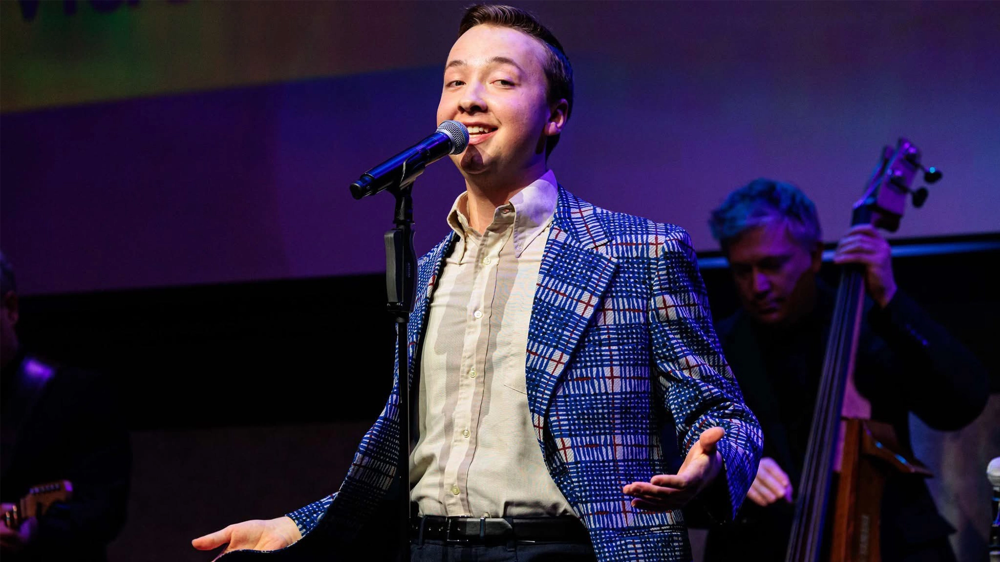 A young man, Josh Thone, sings into a microphone on stage, smiling and wearing a blue patterned blazer over a white shirt. A musician in the background plays an upright bass, with stage lights casting purple and blue tones across the NYC scene.